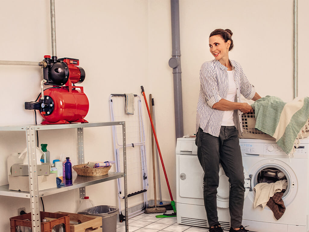 Une femme se tient devant la machine à laver avec son panier à linge et regarde le groupe de surpression Einhell.