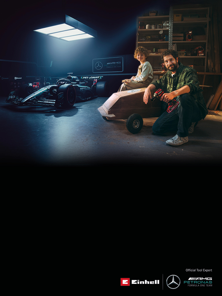 Un père et son enfant dans un atelier avec une voiture à savon faite maison. À l’arrière-plan, une voiture de Formule 1 Mercedes-AMG PETRONAS et des outils Einhell.