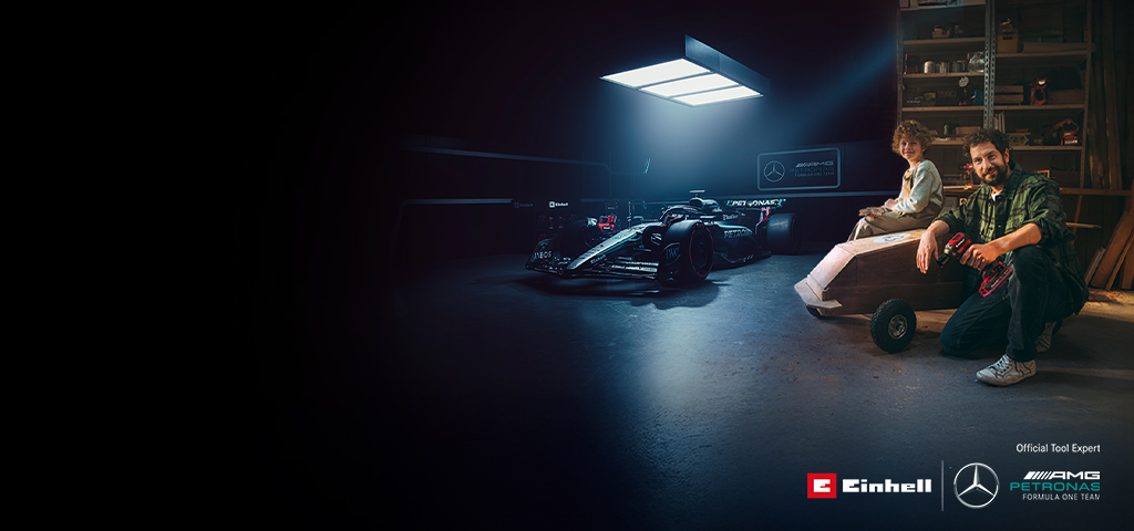 Un père et son enfant dans un atelier avec une voiture à savon faite maison. À l’arrière-plan, une voiture de Formule 1 Mercedes-AMG PETRONAS et des outils Einhell.